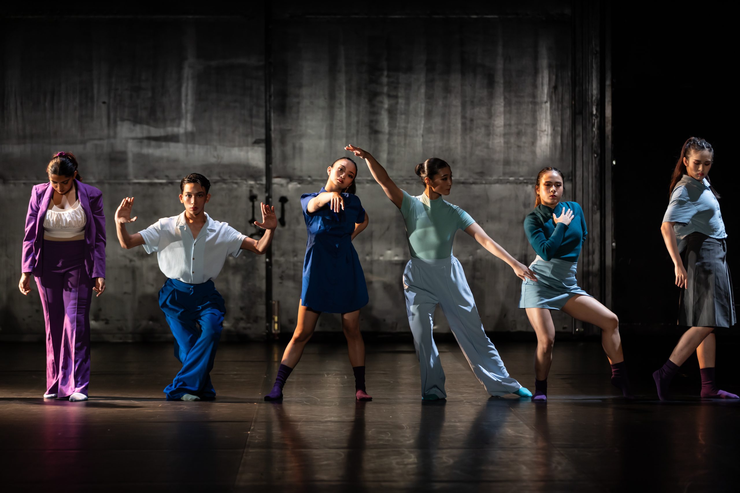 Six dancers in coordinated outfits pose expressively on a dimly lit stage with a textured grey cement wall in the background.