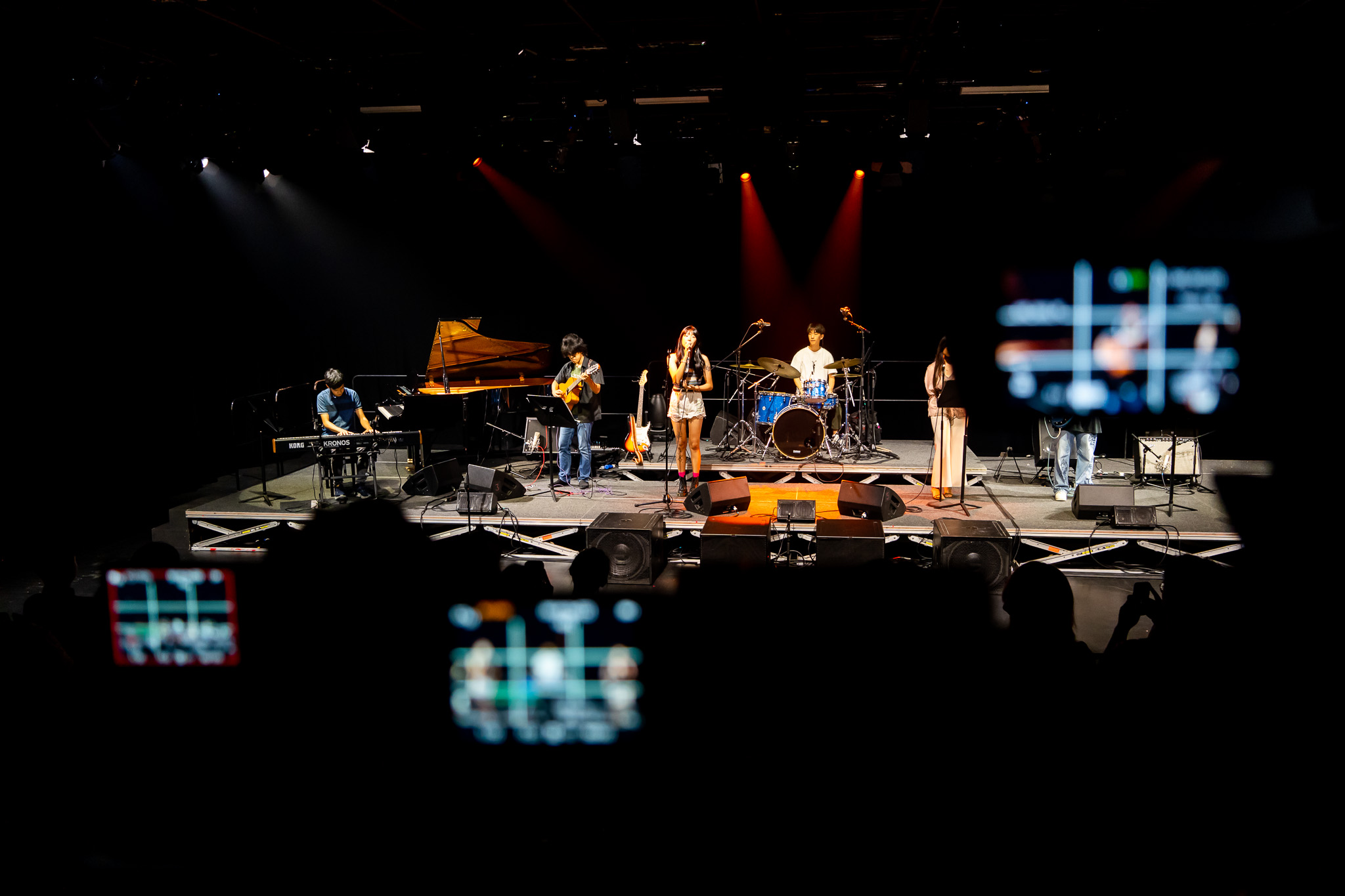 Young musicians perform on a dim stage with phones visible in the foreground, capturing the vocalist, guitarist, drummer and others.