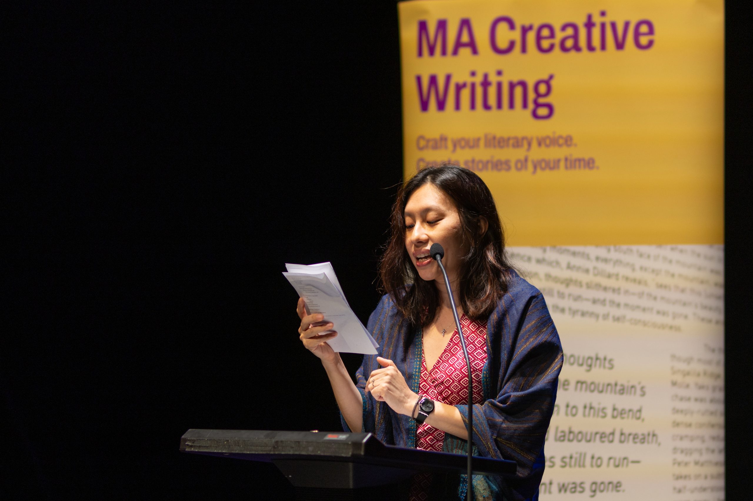 Woman reads from a script at a podium during a literary event, with an MA Creative Writing banner displayed in the background.