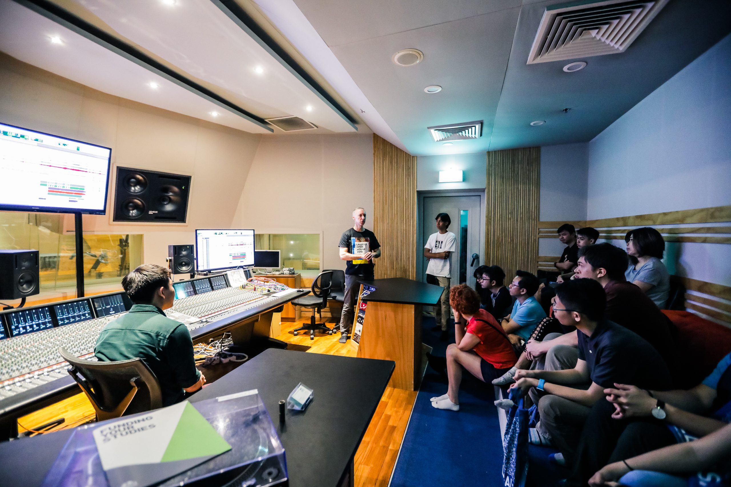 Students in a recording studio watch a lecturer near a mixing console, with audio software on screens and a booth visible through glass.