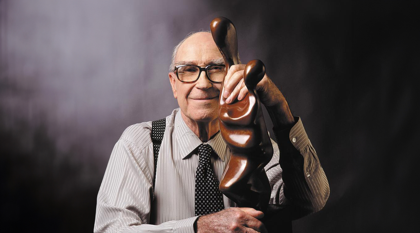 Brother Joseph McNally in striped shirt and tie smiling warmly while resting his head and hand on an abstract wooden sculpture against a dark background.