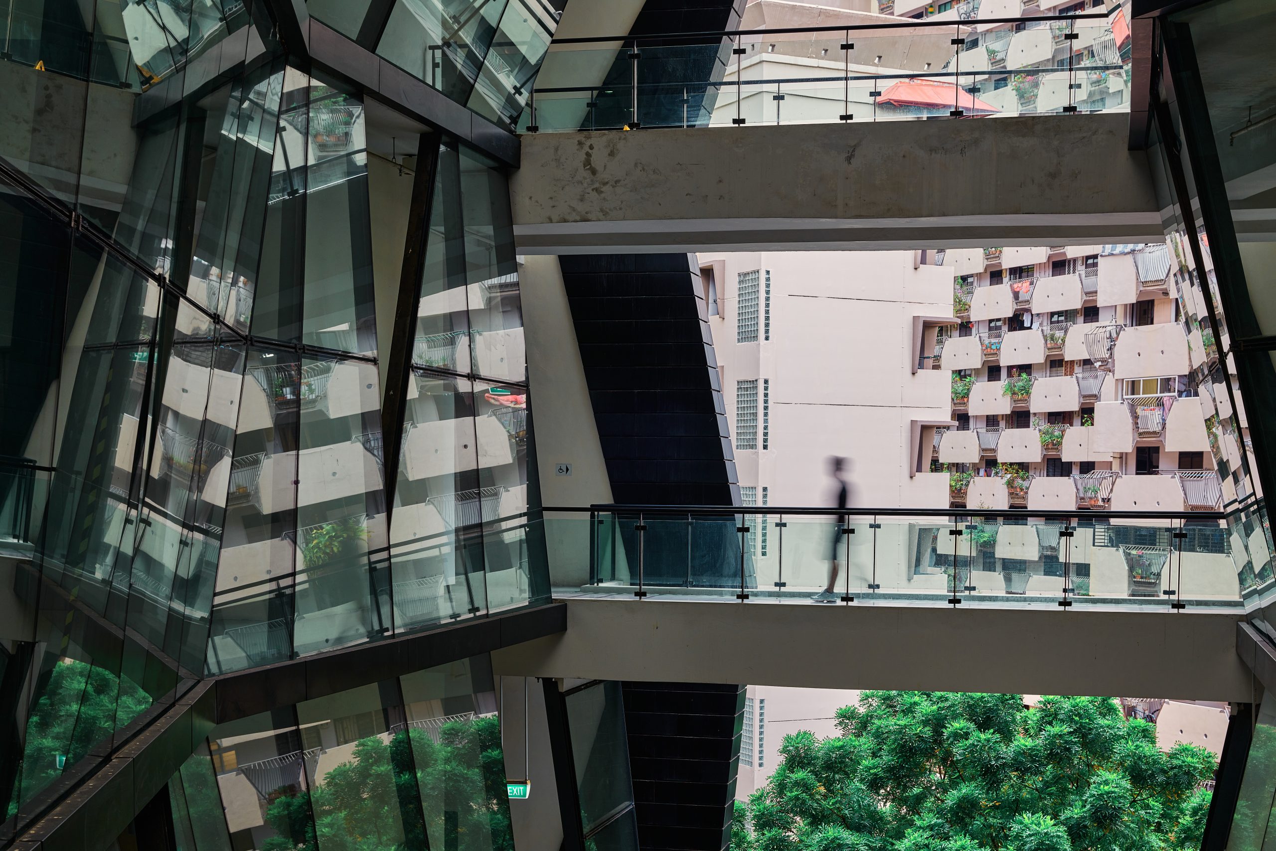 A blurred silhouette of a person walks across LASALLE's glass skybridge inside its angular building, with reflections and geometric shapes juxtaposed against a neighbouring HDB block and lush trees outside.