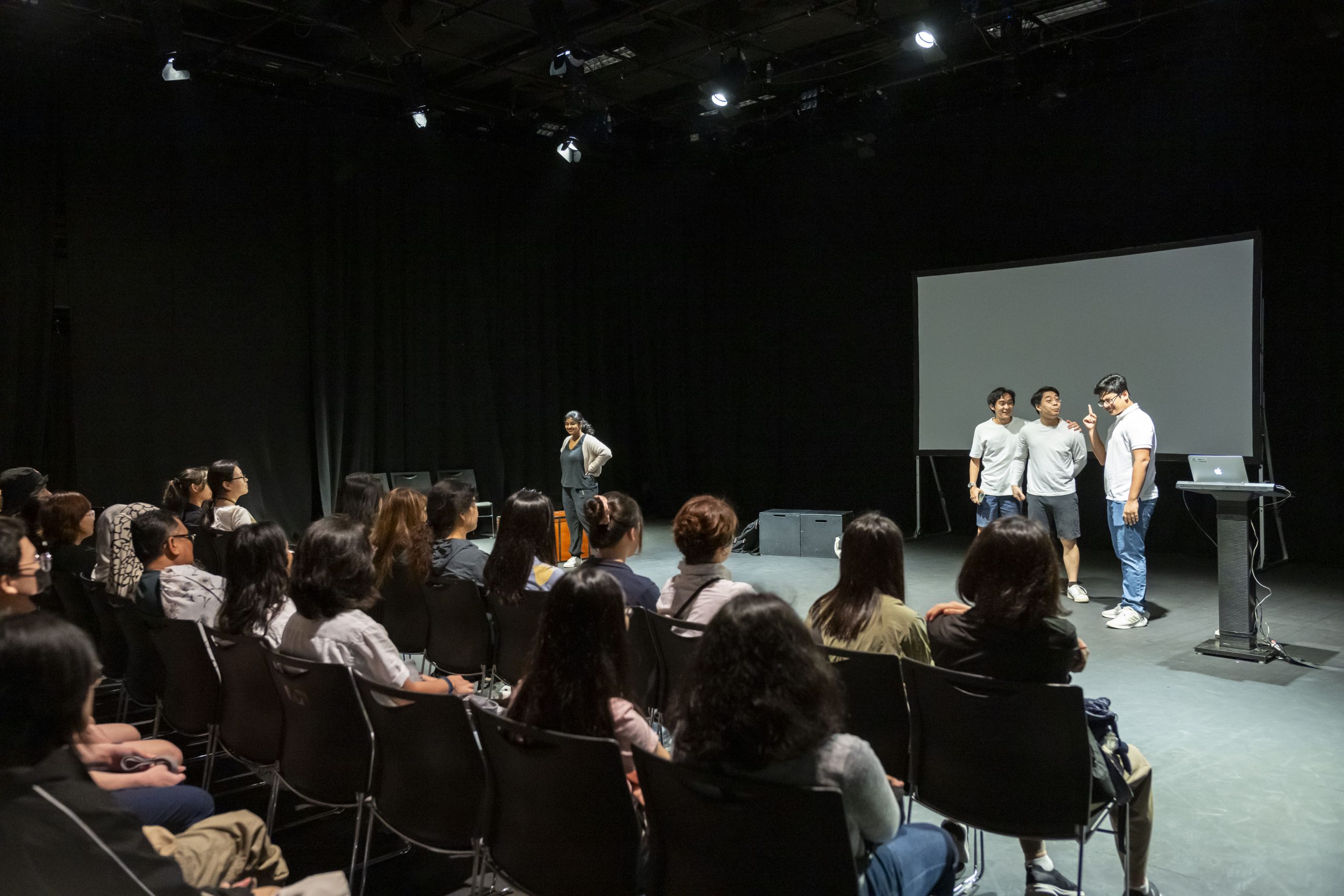 Students perform and present on stage in a black box theatre while an audience observes the session in a dimly lit setting.