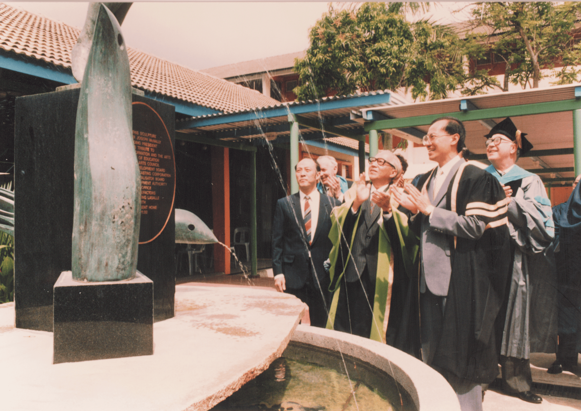 Vintage photography of LASALLE leadership and dignitaries clapping beside a commemorative bronze sculpture fountain on campus, dressed in academic robes and formal attire.