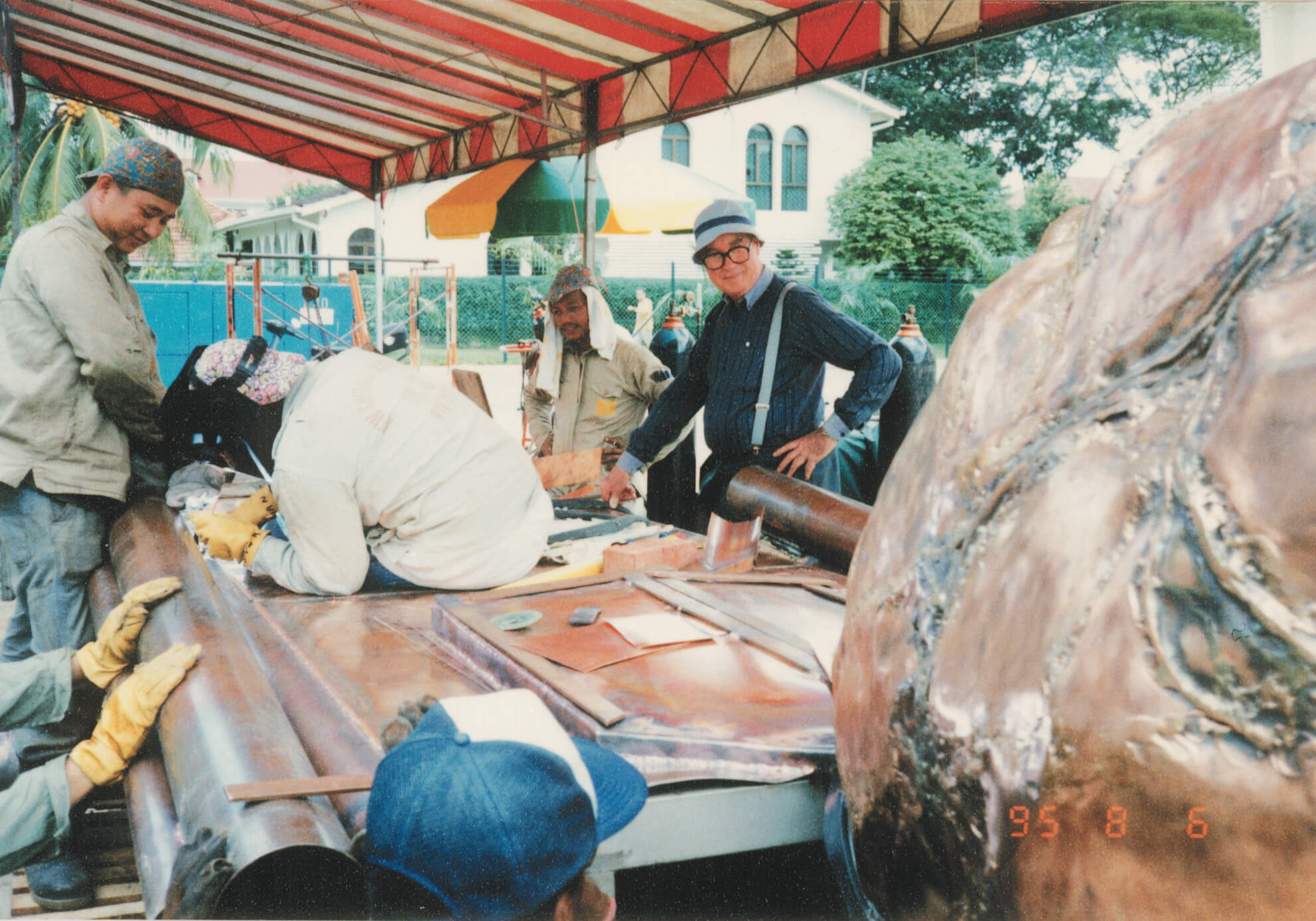 Vintage photography of artists and metalworkers welding and assembling a large outdoor sculpture under a red-and-white canopy, with Brother Joseph McNally observing the process.