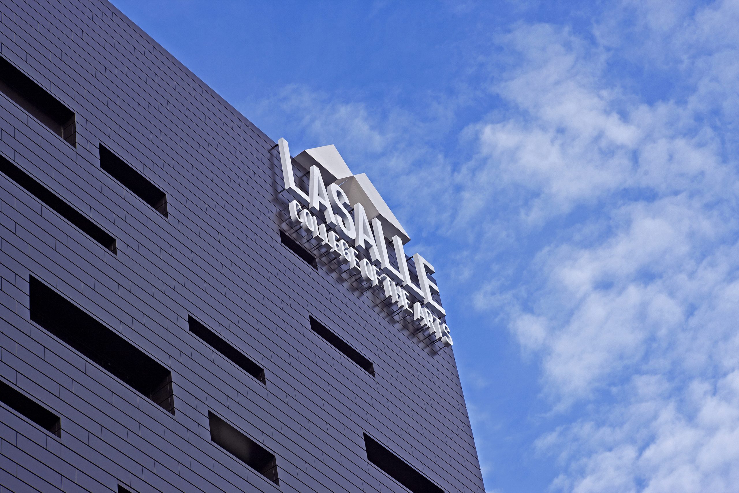 Exterior view of LASALLE College of the Arts building façade with bold signage against a bright blue sky with scattered clouds.