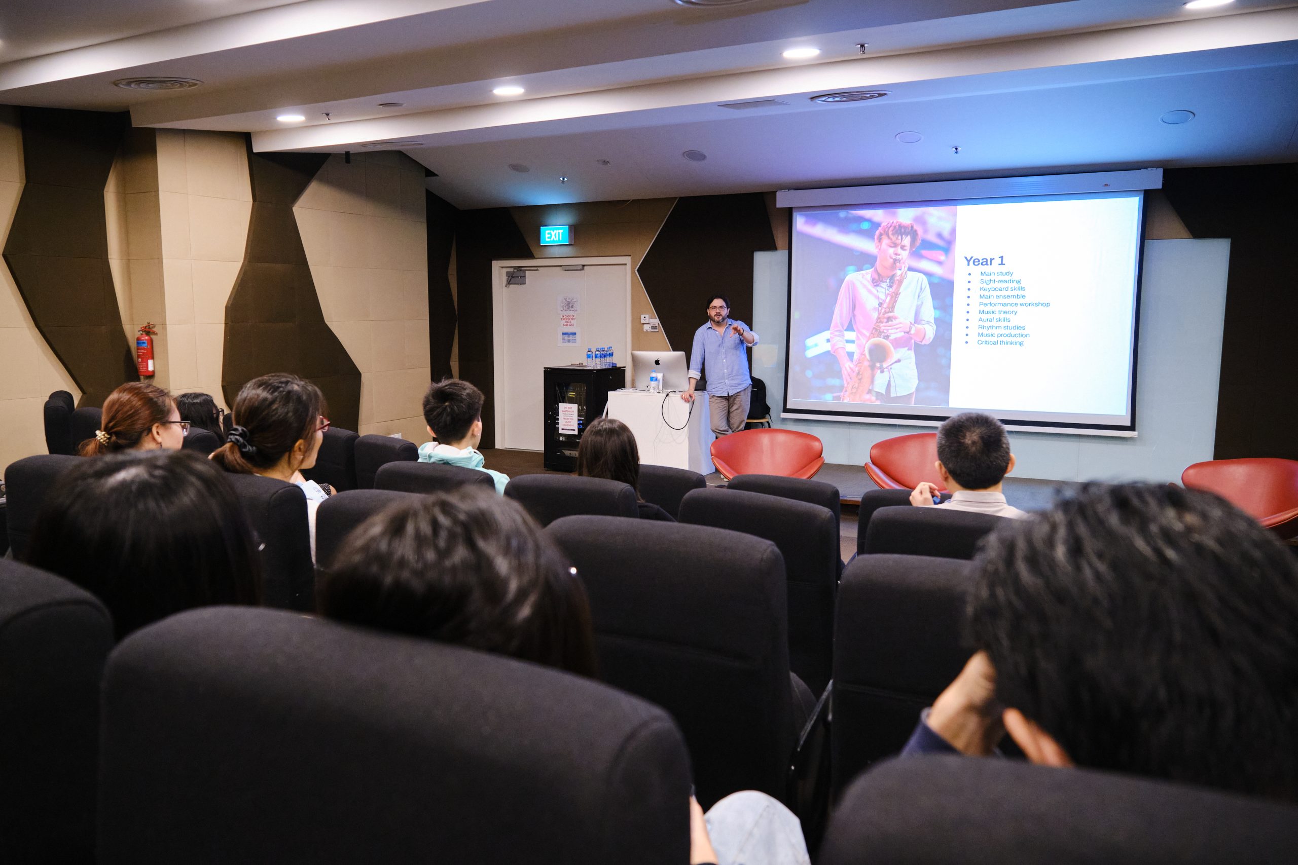 Lecturer presenting to seated students in a darkened room with a large screen showing a musician and course overview.