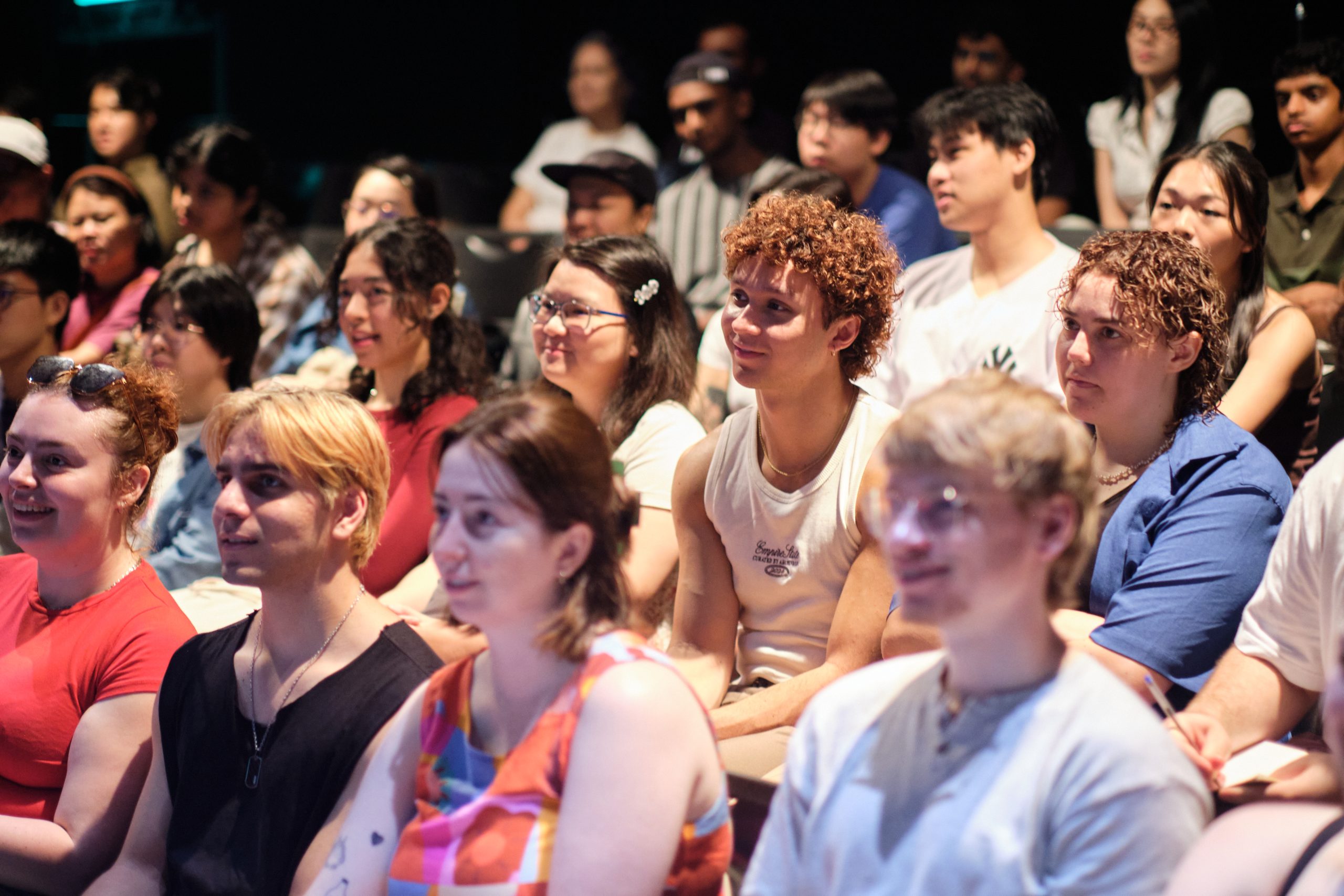 Diverse group of students seated closely together, attentively watching a presentation in a darkened lecture or theatre space.