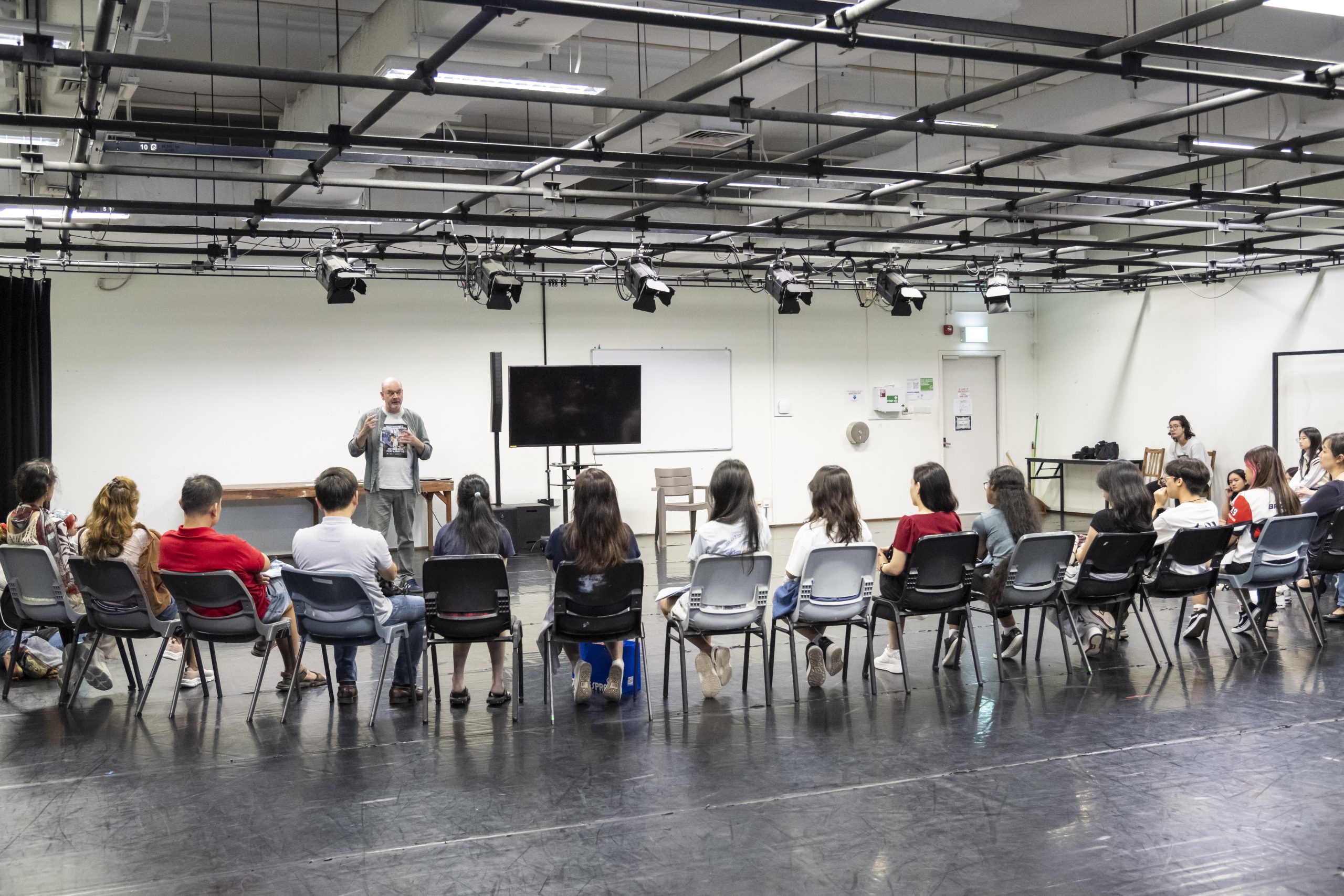 Students seated in a circle listening to a lecturer in a black box studio with stage lighting and a mounted screen.