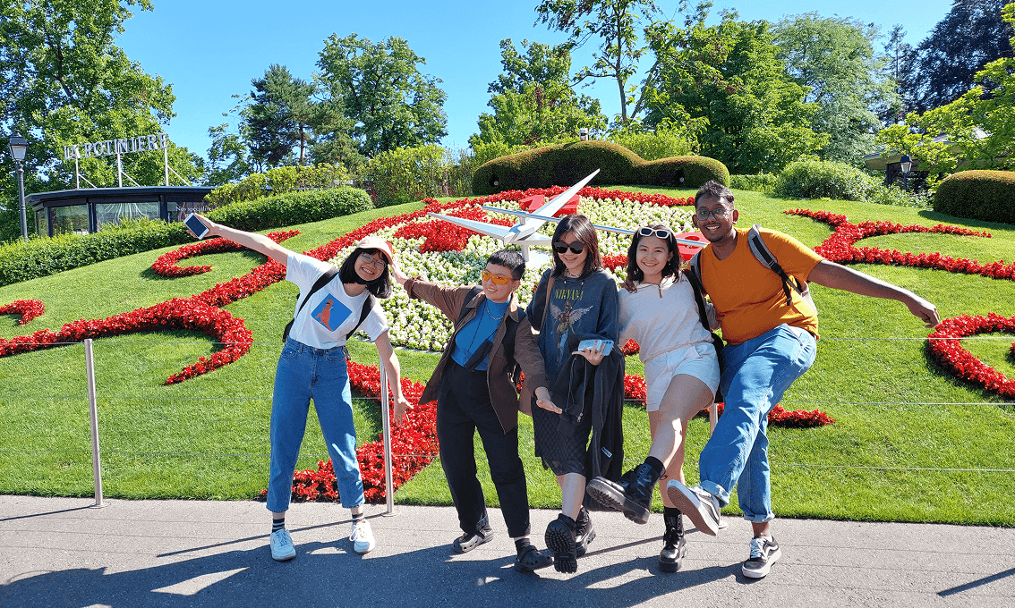 Five LASALLE students posing playfully in front of Geneva’s famous flower clock on a sunny day, with bright red and white floral patterns behind them.