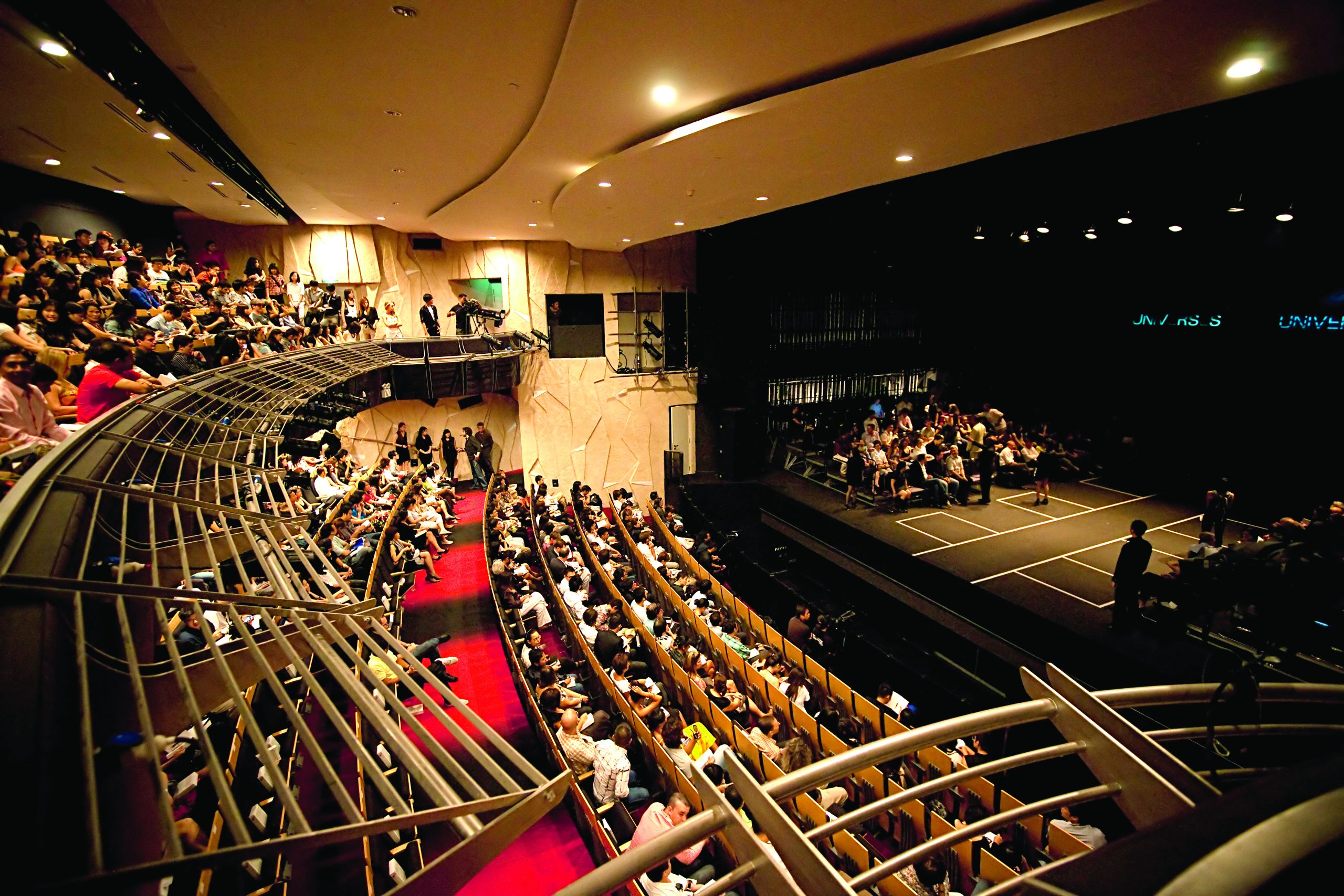 Full audience view inside a modern theatre with curved balconies, red carpet aisle and lit performance stage.