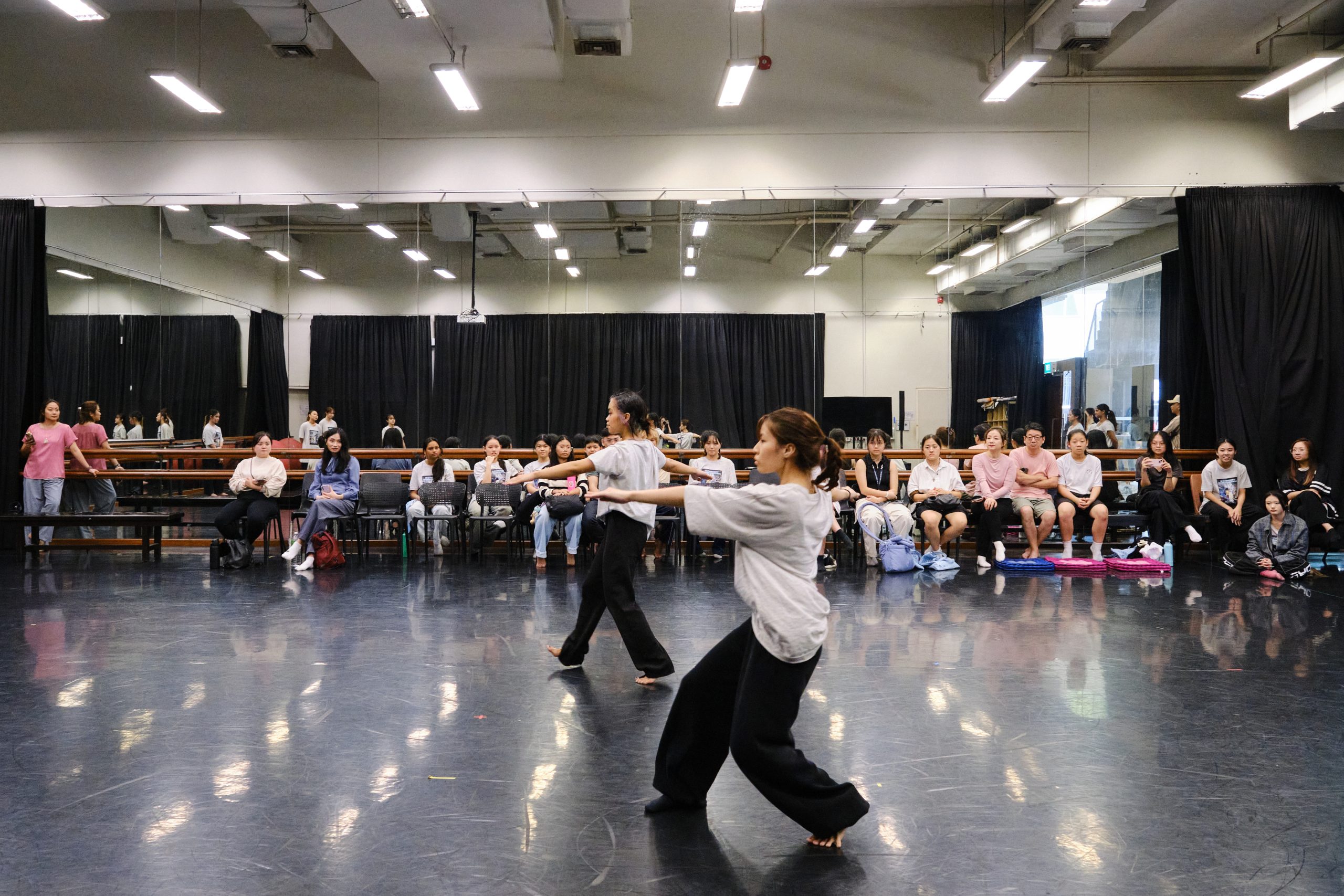 Two LASALLE students performing a contemporary dance piece in a mirrored studio, observed by classmates seated along the back wall.