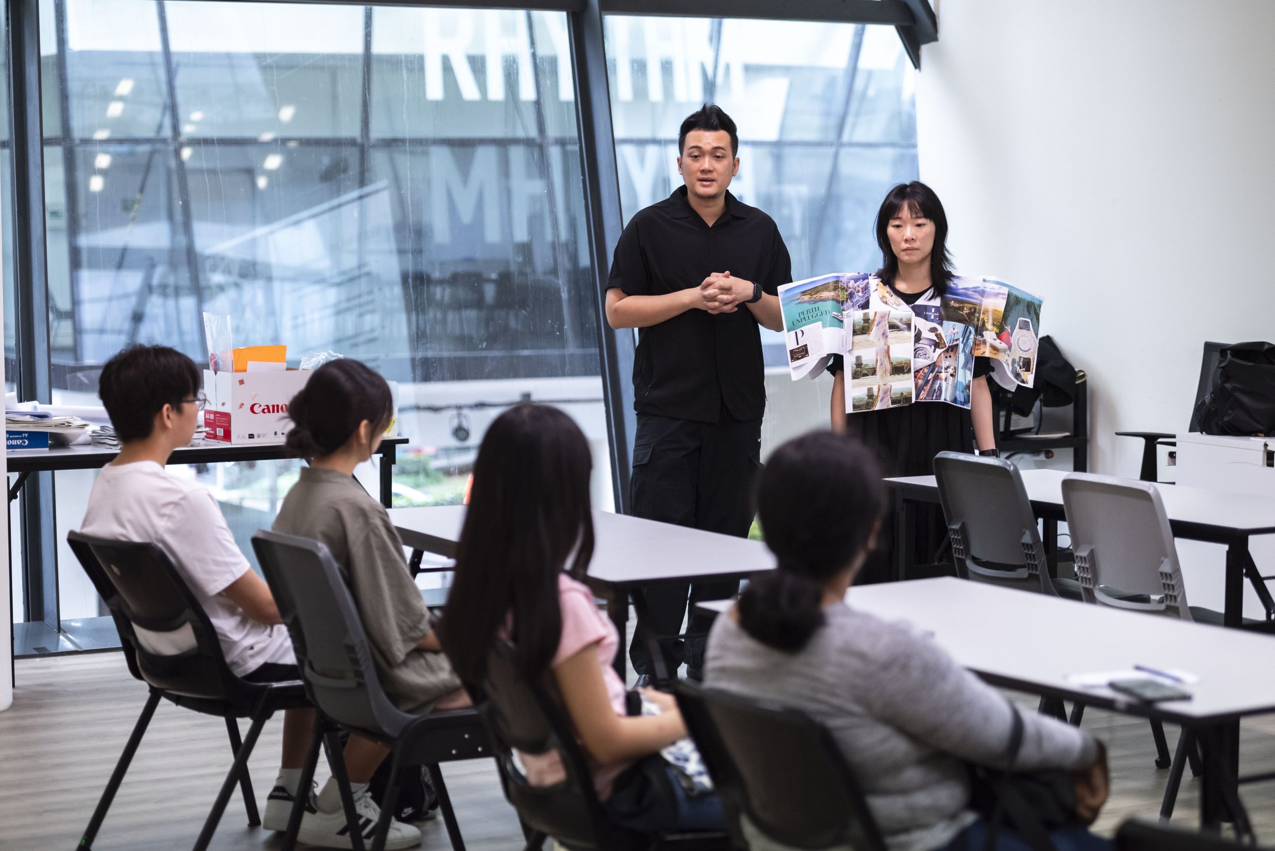 Two presenters speak to seated students in a classroom, with one wearing a creative garment made of printed materials.
