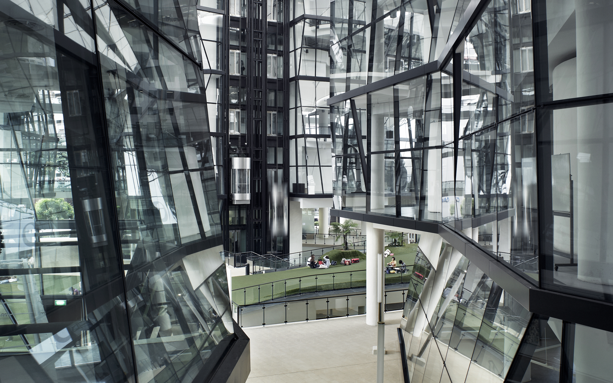 Glass-clad interior of LASALLE’s McNally Campus with lift shafts, angled walkways and students seated on the grass courtyard below.