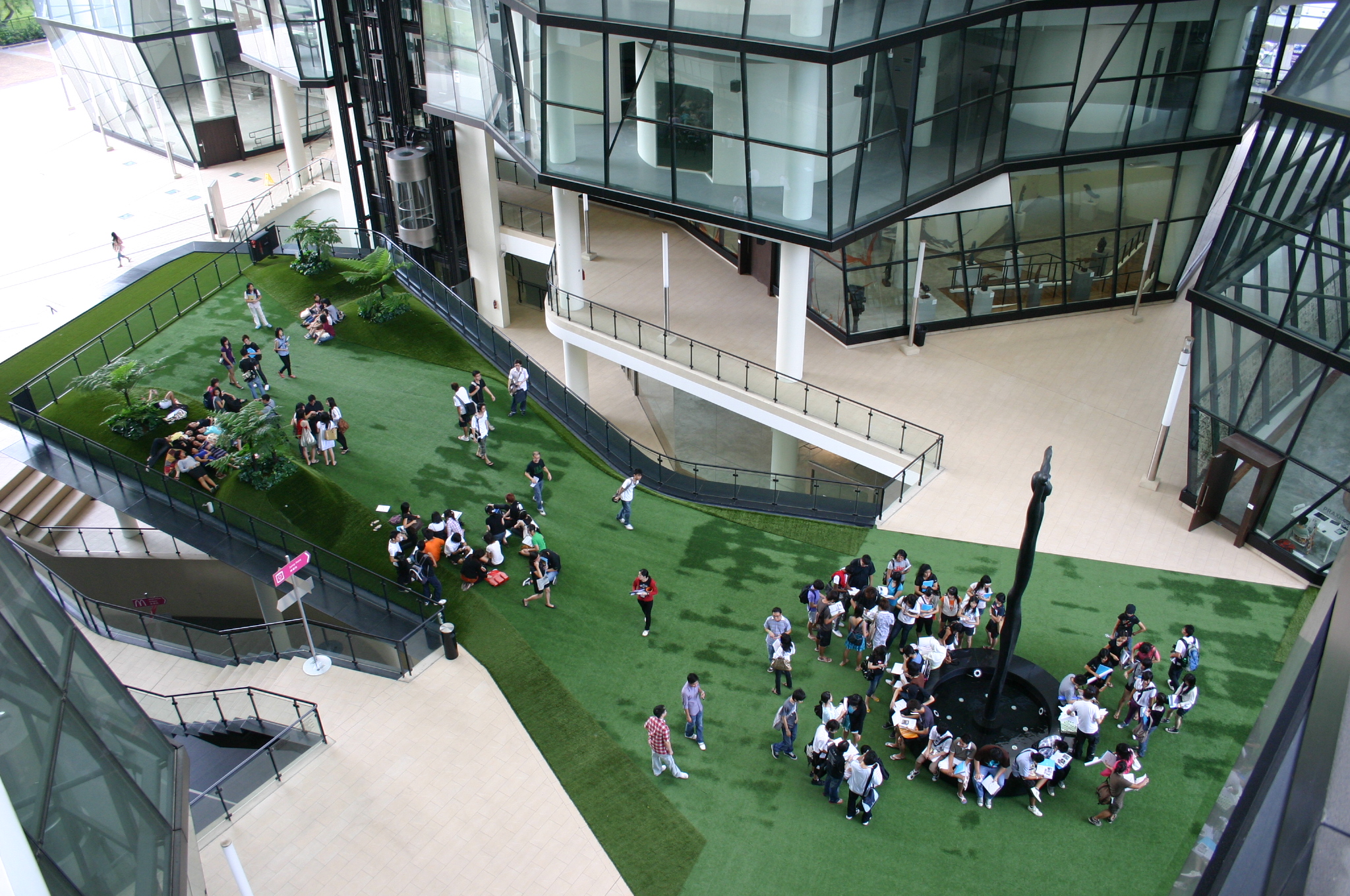 LASALLE students gathering in groups on the green outdoor Campus Green courtyard surrounded by glass buildings and sky bridges.