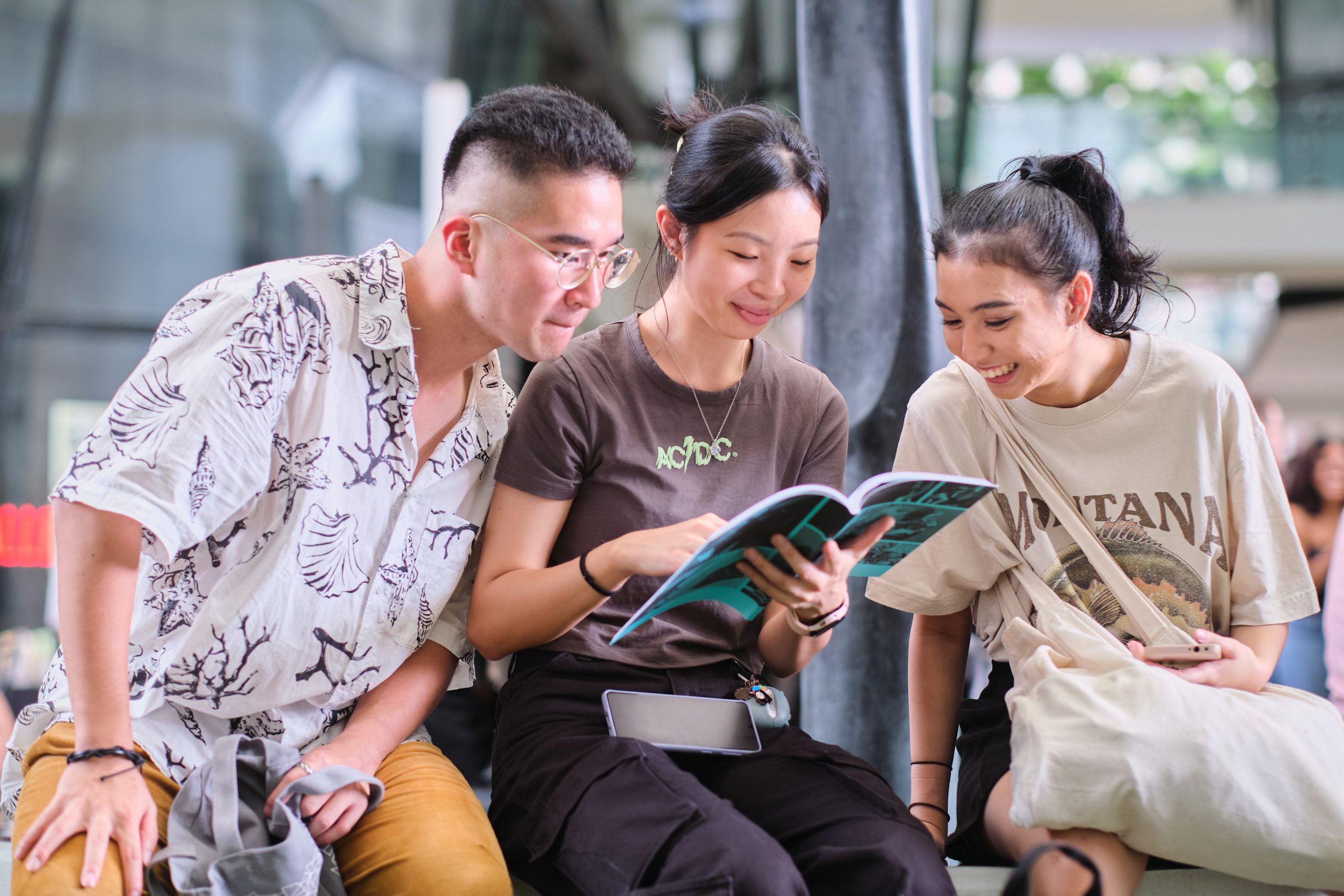 Three students sit together smiling while reading a booklet, wearing casual attire and with campus architecture visible in the background.