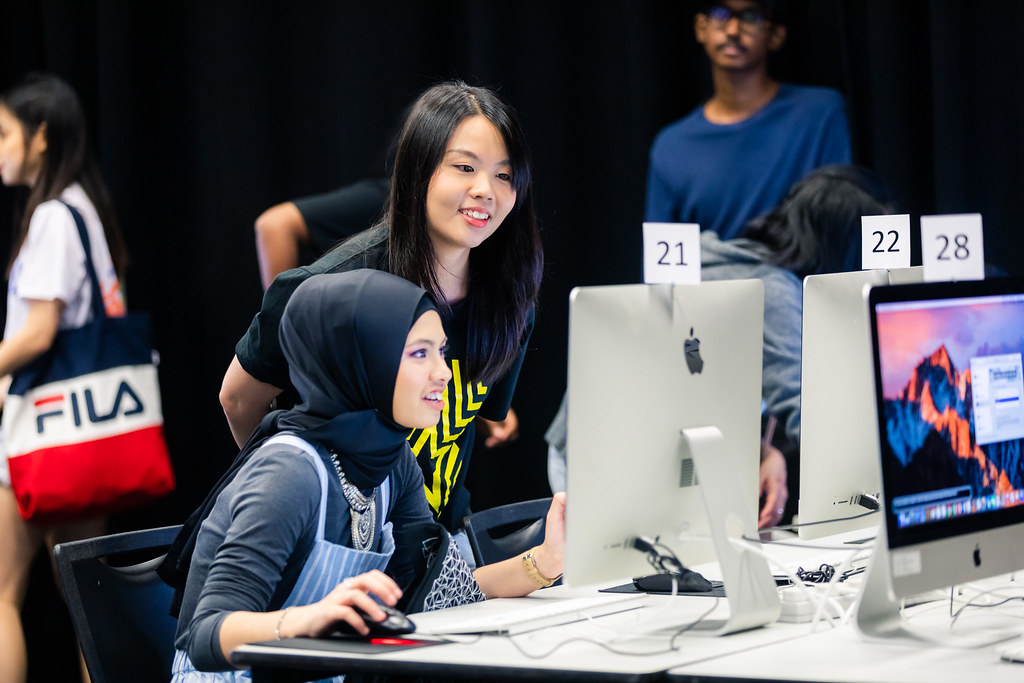 Two students smile as they work together at an iMac in a computer lab, surrounded by numbered desktop stations and classmates.