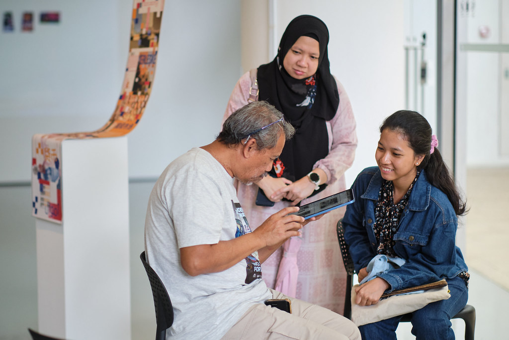 Older man seated showing a tablet screen to two women, one smiling and seated with a tote bag, the other standing and observing attentively