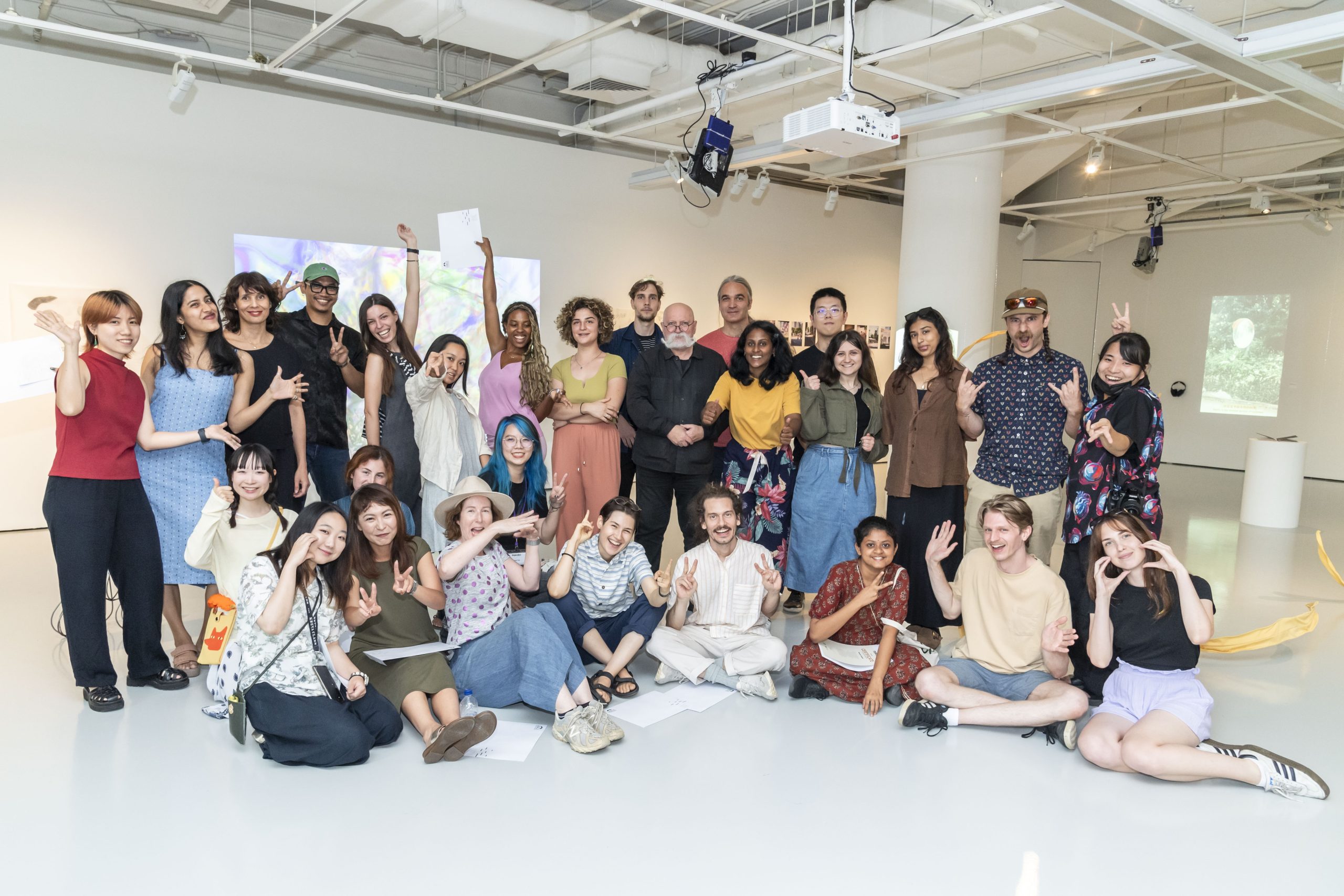 A large group of LASALLE students and staff pose cheerfully together in a gallery space, smiling and making playful gestures.