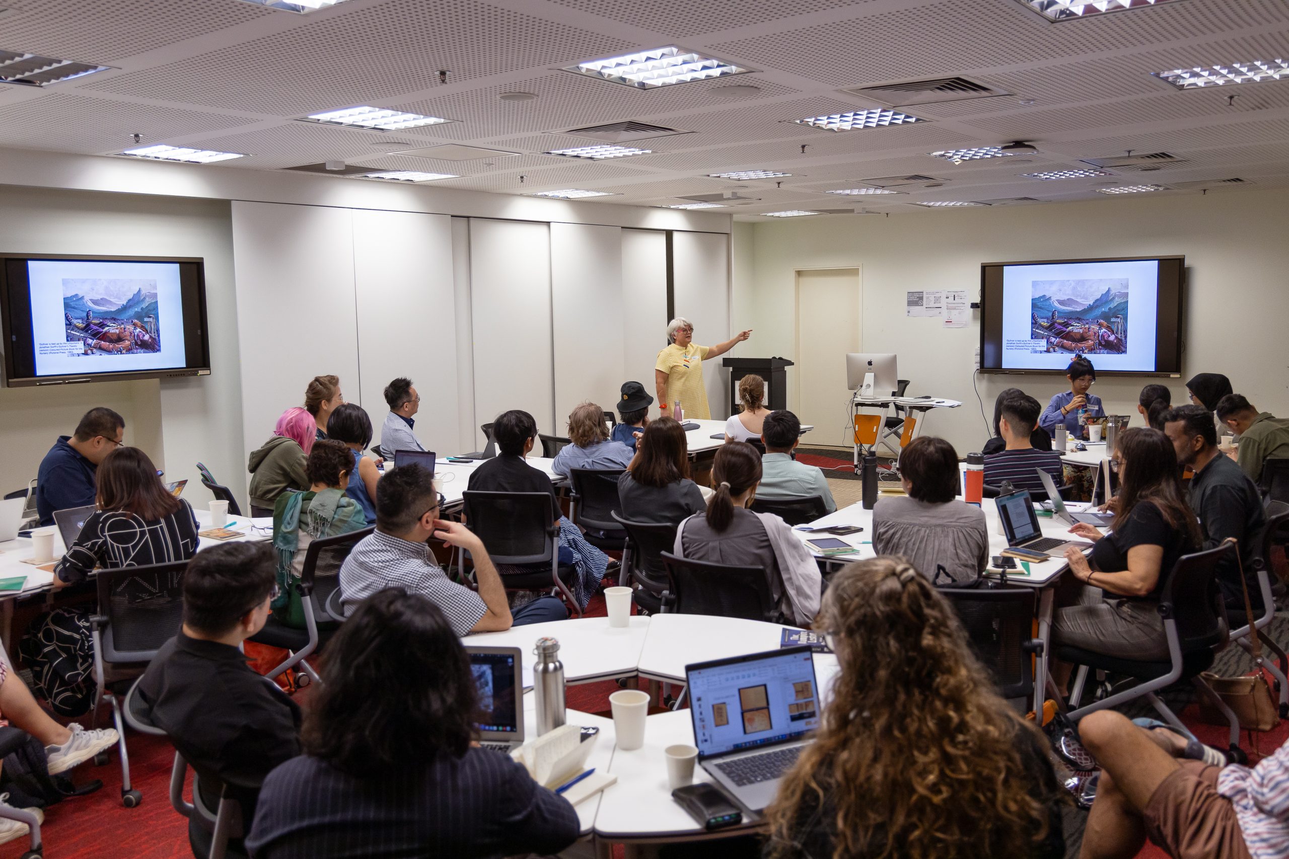 A lecturer presents to a full classroom at LASALLE, with students seated at tables using laptops and slides projected on screens.