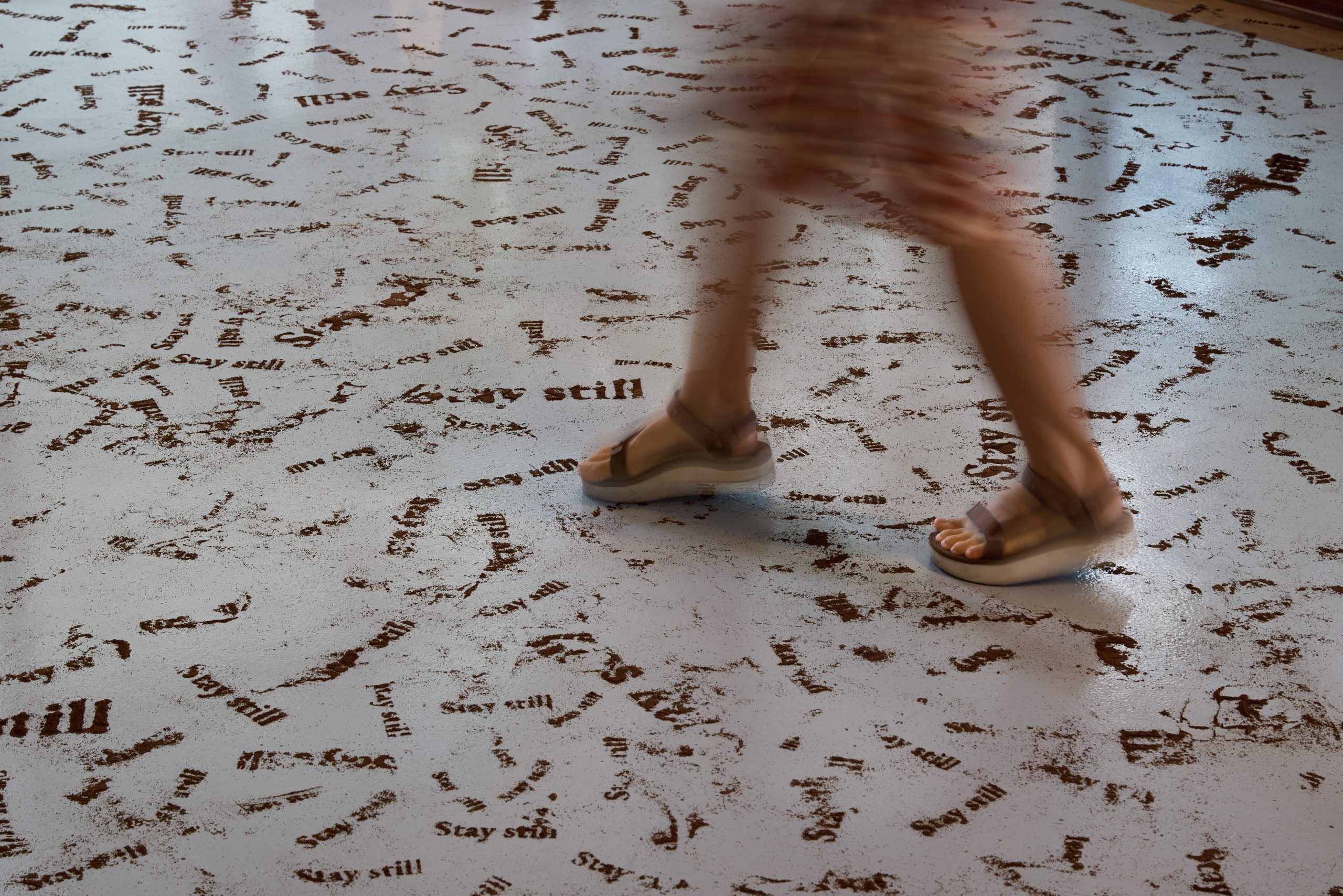 Close-up of feet walking across a gallery floor covered in repeated stamped words “Stay still” forming part of an installation.