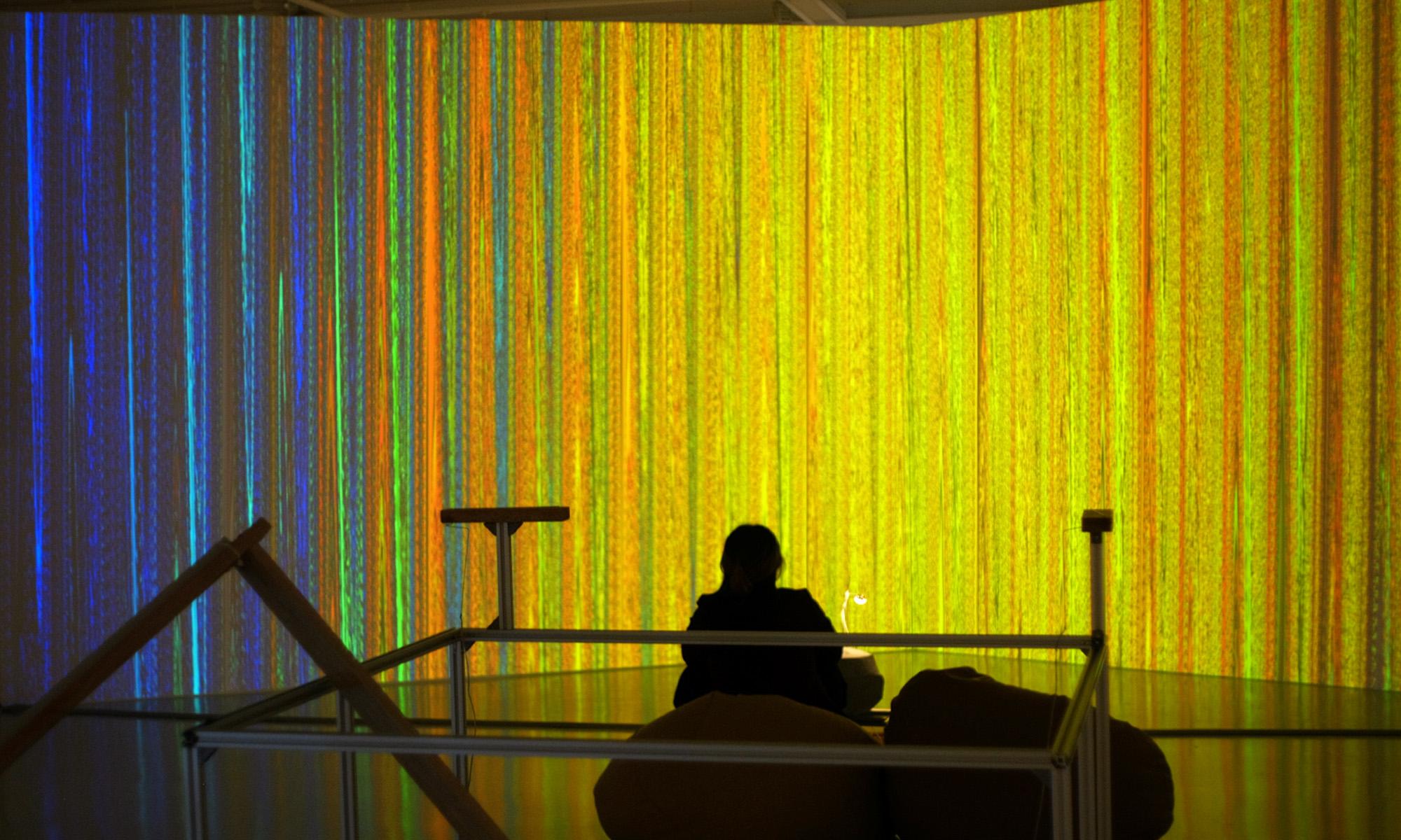 A visitor sits before a LASALLE installation with floor-to-ceiling projections of shifting vertical light patterns in blue, orange and yellow.