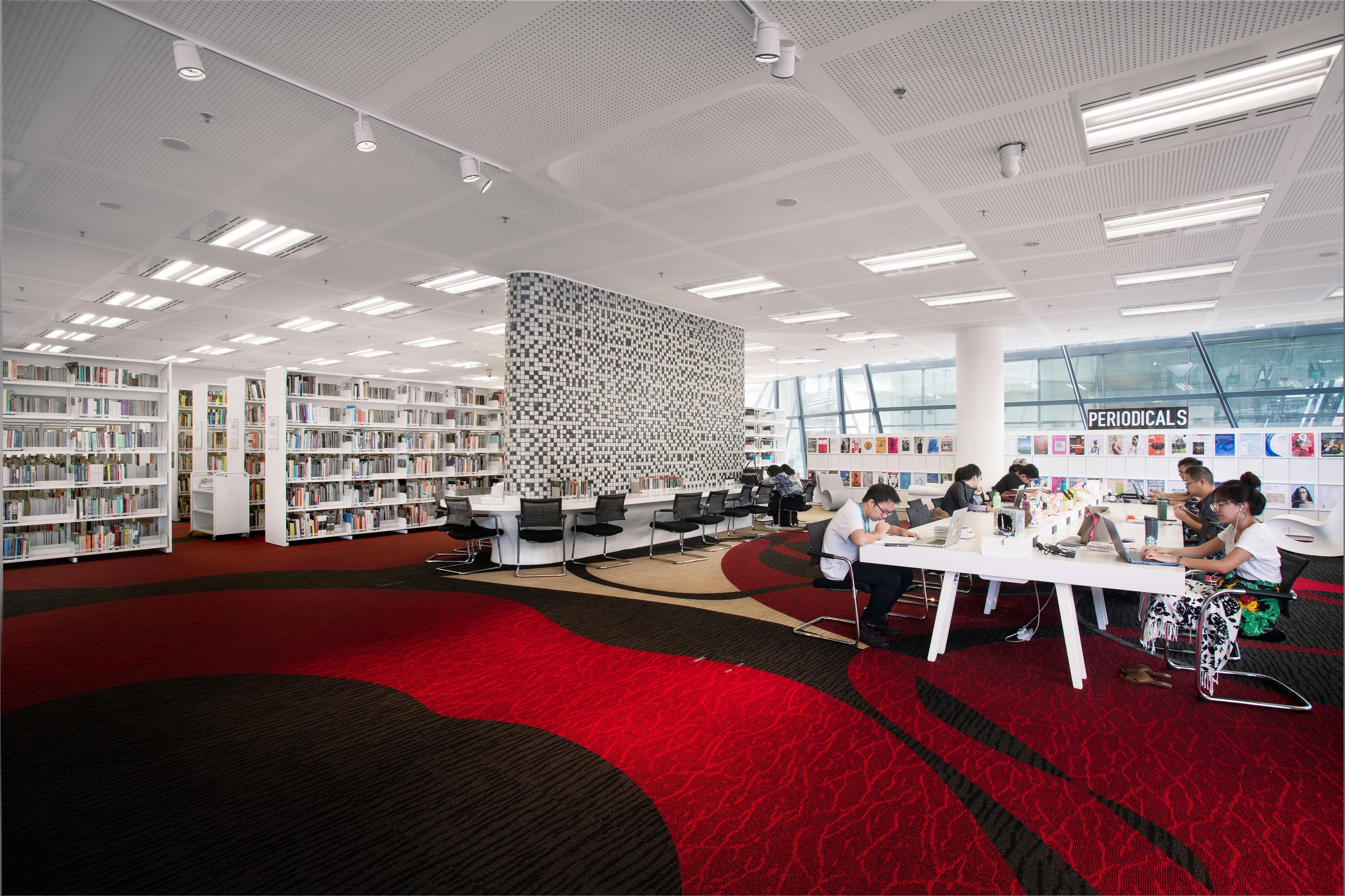 Students study at long tables in a spacious campus library with red-patterned carpets, bookshelves and natural light from tall windows.