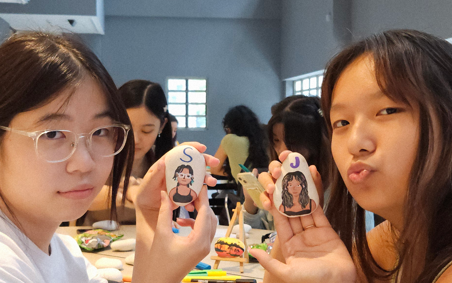 Two students show off painted character portraits on stones, with others in the background participating in the creative workshop.