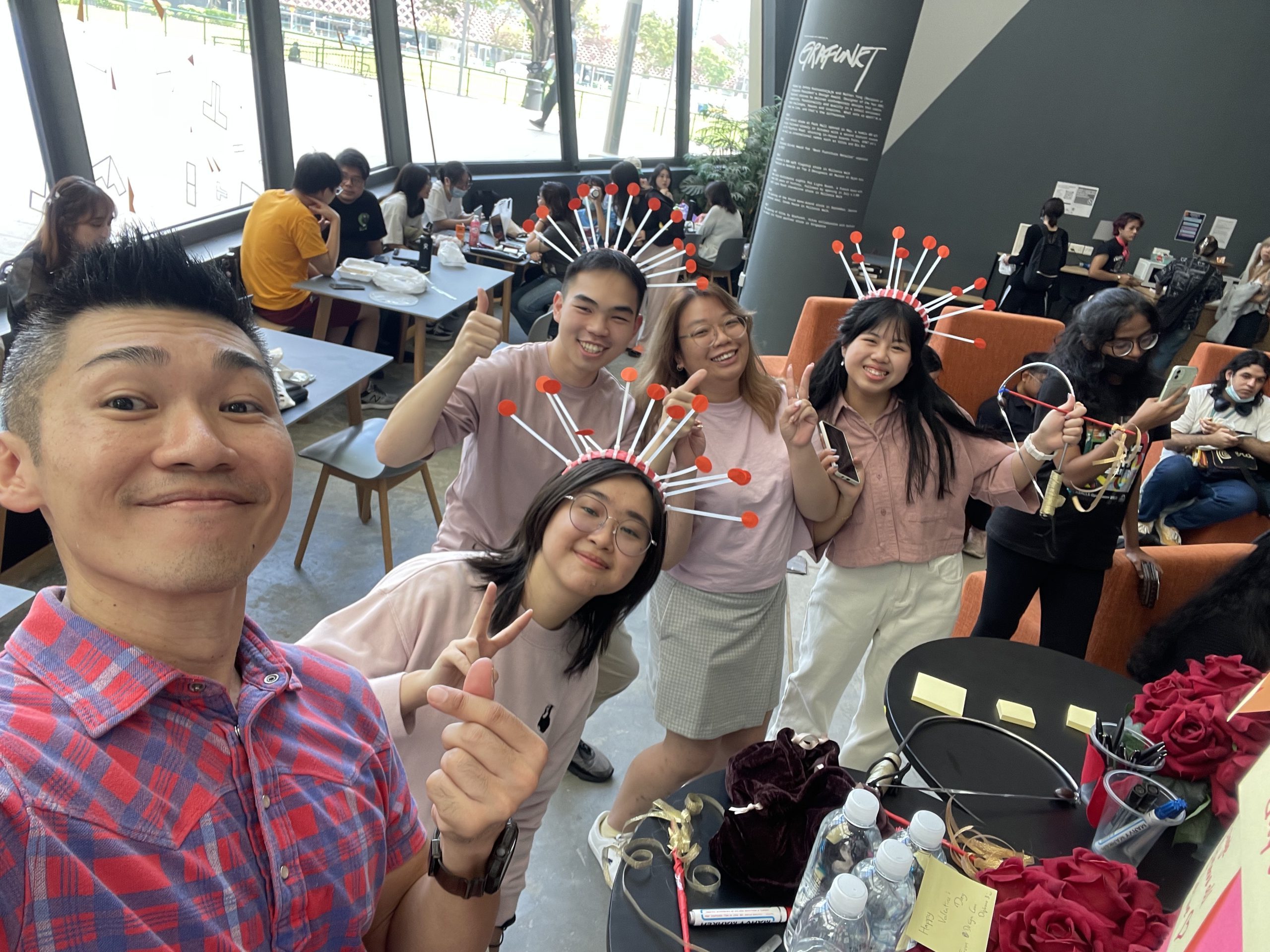 LASALLE students pose in a café space wearing handmade headgear with red-tipped antennae, surrounded by crafts and activity props.