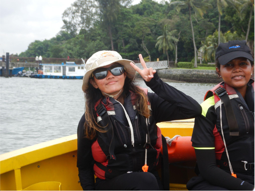 Two women in life vests and caps on a yellow boat, with one flashing a peace sign, against a tropical island backdrop.
