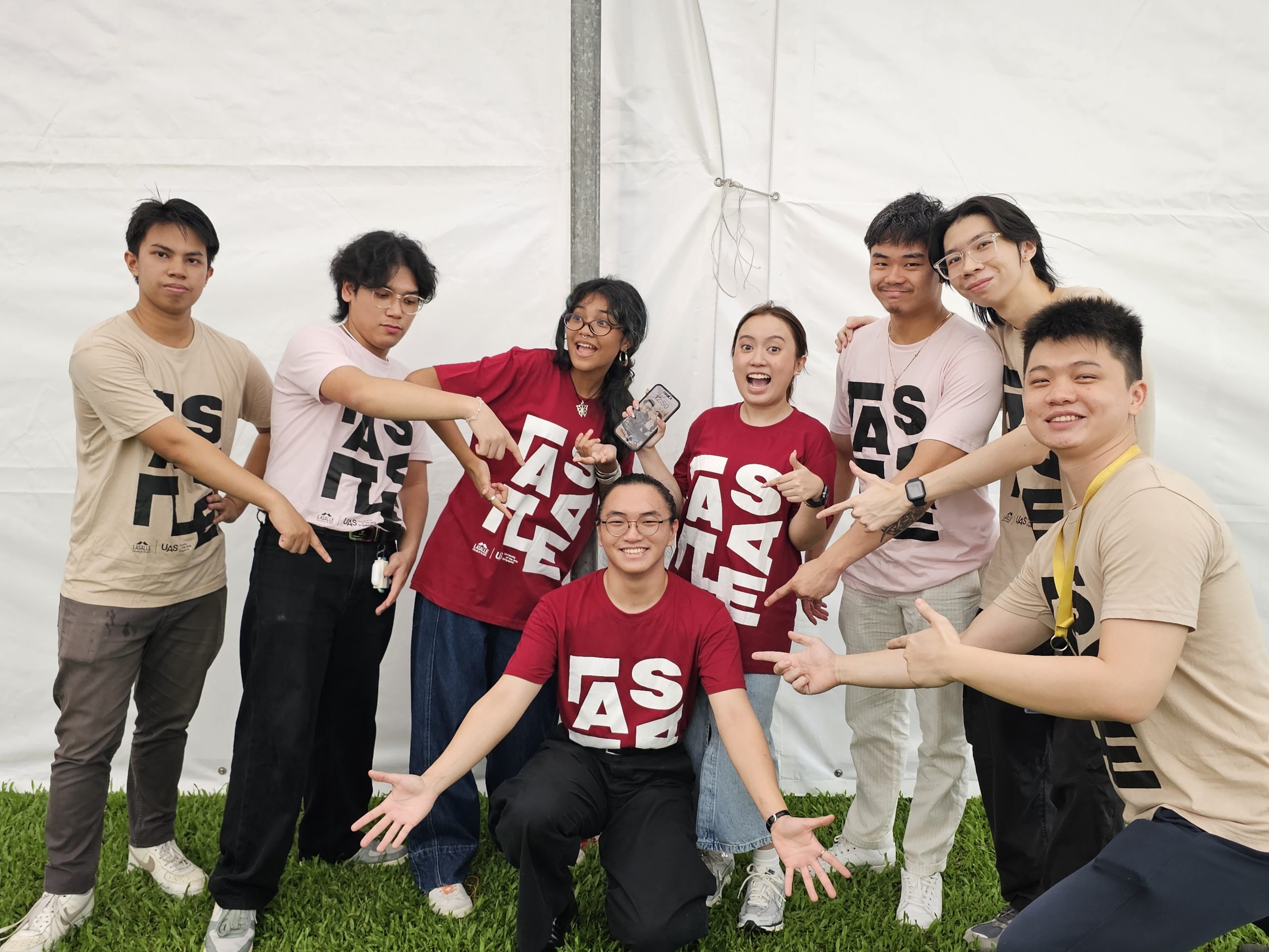 Group of students in LASALLE t-shirts posing playfully in front of a white tent, pointing at two peers in red shirts.