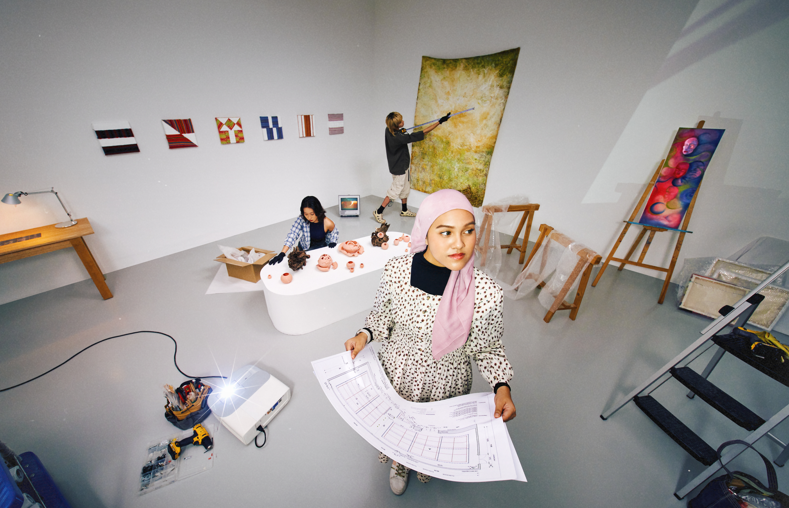 Three students setting up an exhibition space with ceramics, paintings and tools, while one foreground figure in a pink headscarf holds architectural floor plans.