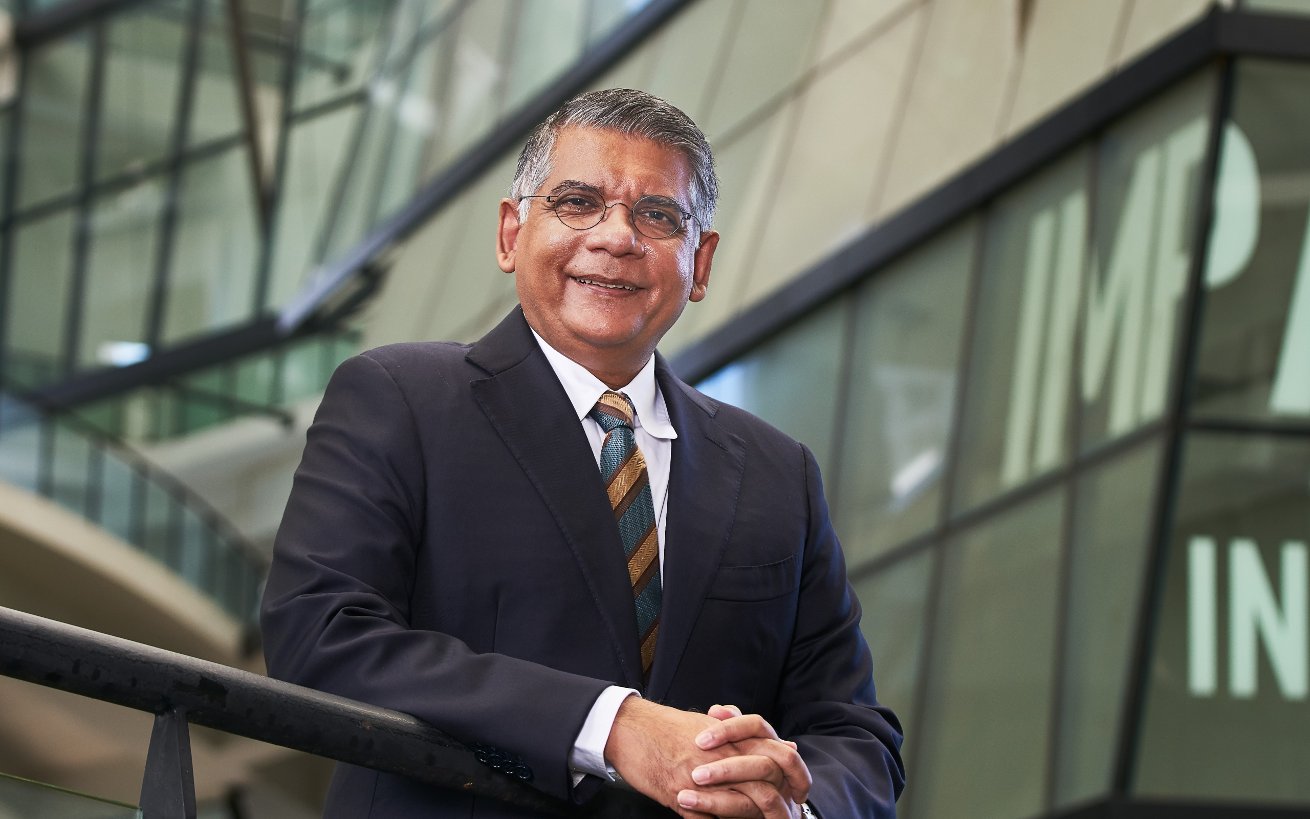 Man in a dark suit and striped tie smiling with hands clasped, standing in front of a modern glass building with the word “IMPACT” partially visible.